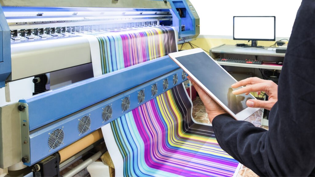 A large-format printer producing a vibrant, multicolored print while a person in a black suit monitors the process using a tablet